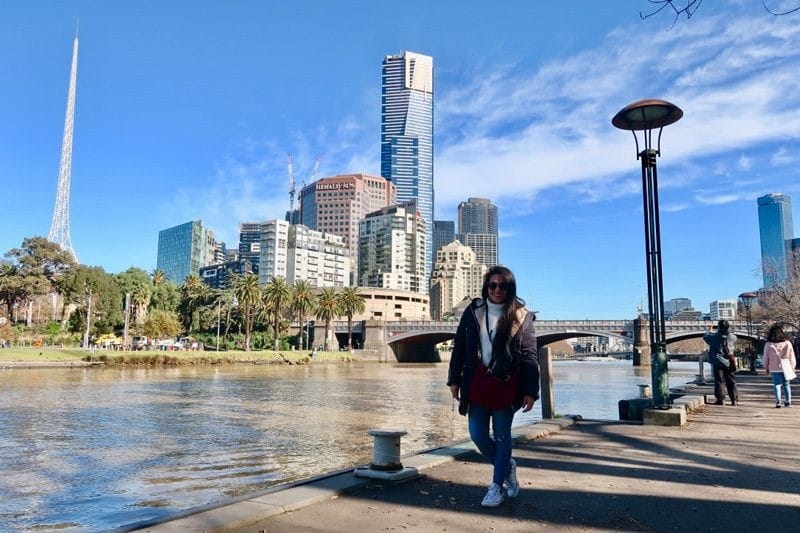 Ciudad de Brisbane chica morena con abrigo y bambas posando delante de la ciudad de Brisbane en Australia. Detrás de ella hay el río con un puente y los rascacielos de la ciudad
